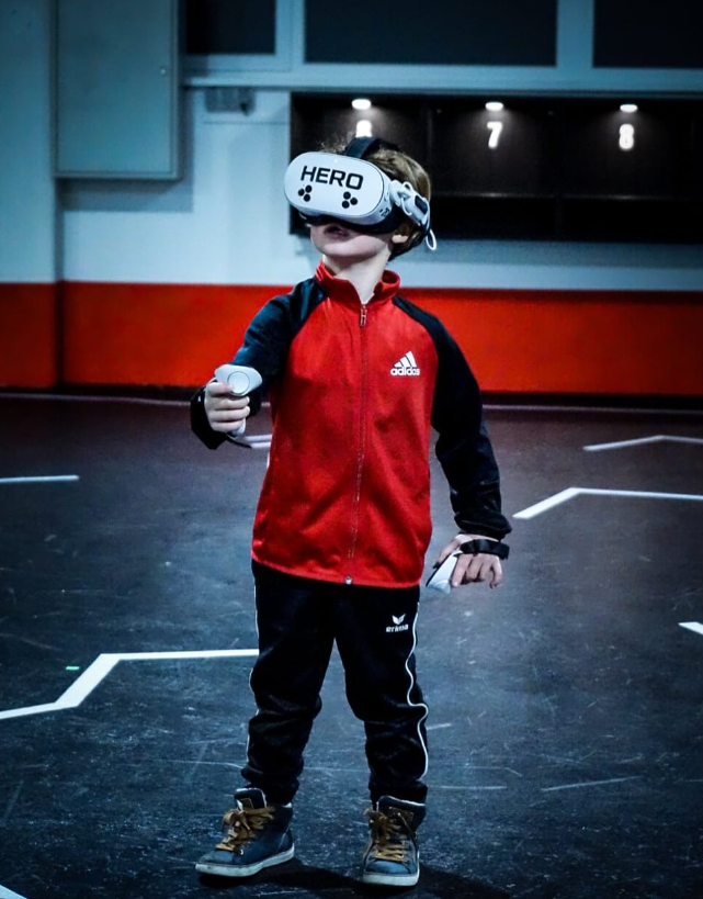 Kind im roten Trainingsanzug mit VR-Brille und zwei Controllern in der VR-Arena